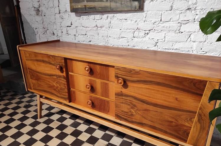 Drewniany kredens w stylu vintage z widocznym fornirem o bogatym usłojeniu, ustawiony na tle białej ceglanej ściany i podłogi w szachownicę.
