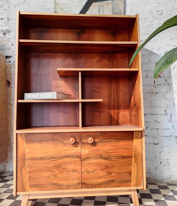 Regał z drewna o unikalnym usłojeniu, w stylu vintage, z półkami i szafką z okrągłymi uchwytami, na tle ceglanej ściany i szachownicy na podłodze, z książką 'HOME BOOK' na jednej z półek.