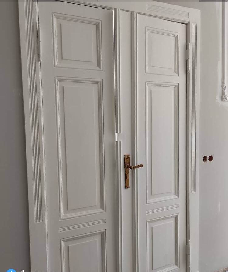 Białe dwuskrzydłowe drzwi wewnętrzne z frezowanymi panelami i mosiężną klamką, widoczne zawiasy i fragment ściany z niedomalowanymi otworami po gniazdkach elektrycznych.