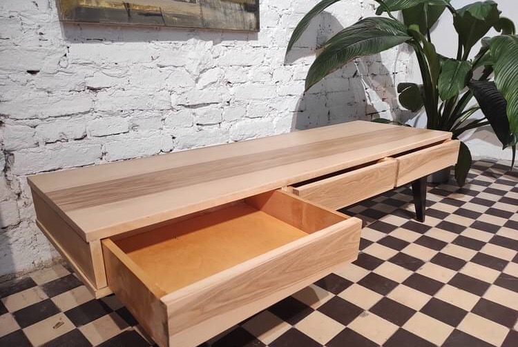 Nowoczesna komoda z jasnego drewna z wysuwanymi szufladami, na tle białej ceglanej ściany i rośliny doniczkowej, ustawiona na podłodze w szachownicę.