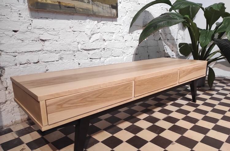 Niska komoda z jasnego drewna z trzema szufladami, ustawiona na czarnych nóżkach, na tle białej ceglanej ściany i rośliny doniczkowej, na podłodze w szachownicę.