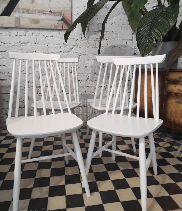 Cztery białe, drewniane krzesła w stylu skandynawskim na czarno-białej podłodze w szachownicę, ustawione na tle białej ceglanej ściany i dużej rośliny doniczkowej.