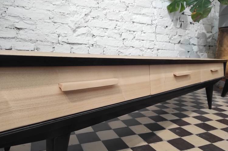Niska komoda z jasnego drewna z czarnymi elementami, widoczne trzy szuflady z minimalistycznymi uchwytami, na tle białej ceglanej ściany i czarno-białej podłogi w szachownicę.