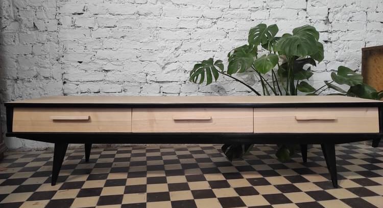 Niska komoda z trzema szufladami z jasnego drewna, czarnymi nogami w stylu retro, na tle białej ceglanej ściany i rośliny doniczkowej, ustawiona na podłodze w szachownicę.