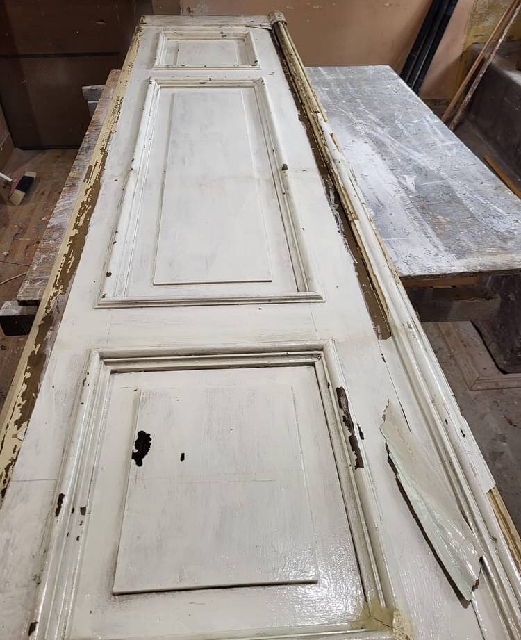 Stara, drewniana, biała drzwi z ubytkami farby i uszkodzeniami, leżąca na stole w warsztacie, w trakcie renowacji.