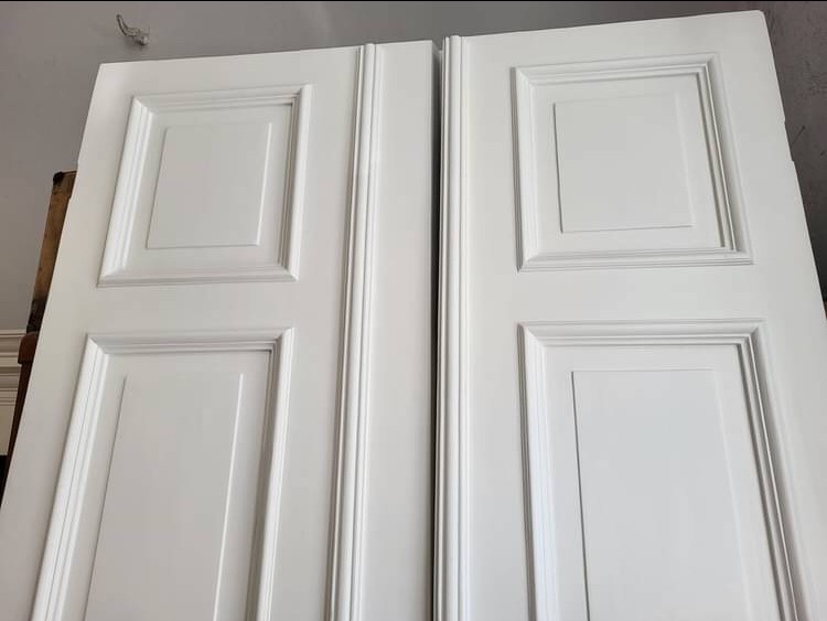 Dwa białe, klasyczne fronty meblowe z płycinami, ustawione obok siebie, oświetlone z góry.