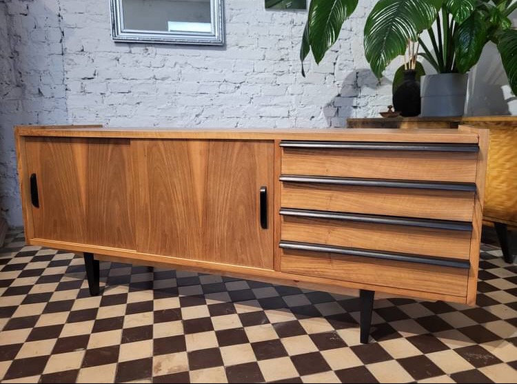 Drewniana komoda w stylu vintage z przesuwnymi drzwiami i trzema szufladami z czarnymi uchwytami, ustawiona na czarno-białej szachownicy, z rośliną doniczkową w tle.