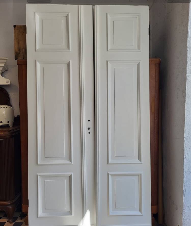 Białe, dwuskrzydłowe drzwi z panelami, opierające się o meble w tle, widoczne ślady po montażu zamka.