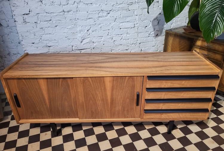 Drewniana komoda z przesuwanymi drzwiami i czterema szufladami z czarnymi uchwytami, na metalowych nóżkach, na tle białej ceglanej ściany i szachownicy na podłodze.