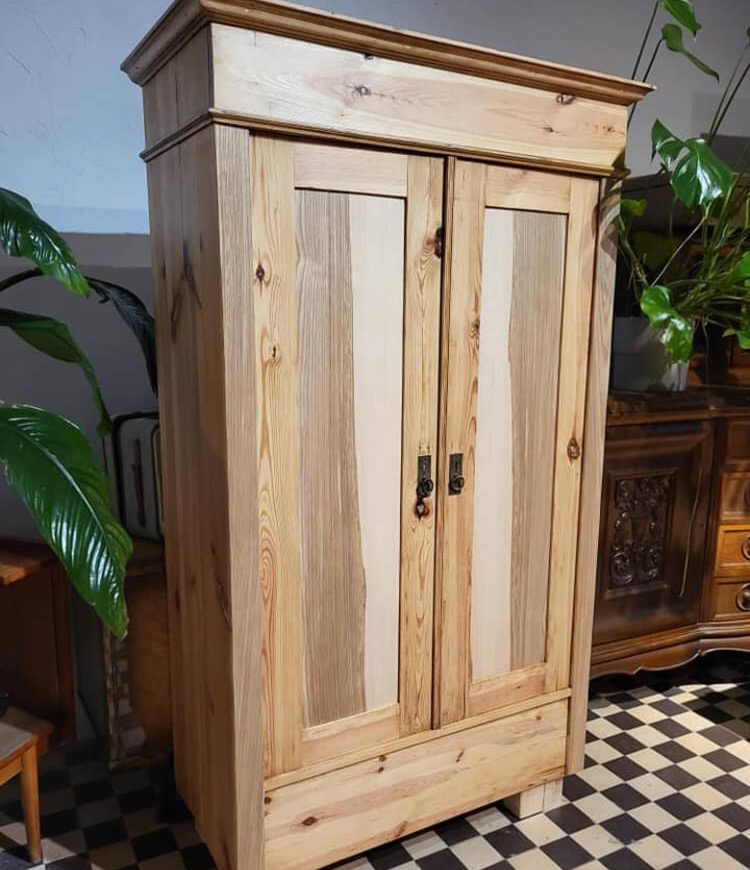 Drewniana szafa dwudrzwiowa z szufladą, wykonana z jasnego drewna z widocznymi słojami, z ciemnymi metalowymi uchwytami, stojąca na czarno-białej podłodze w szachownicę, z roślinami doniczkowymi...