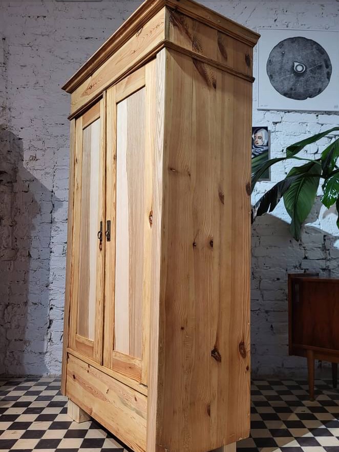 Drewniana szafa dwudrzwiowa z szufladą, wykonana z jasnego drewna sosnowego, z widocznymi sękami, na tle białej ceglanej ściany i czarno-białej podłogi w szachownicę.