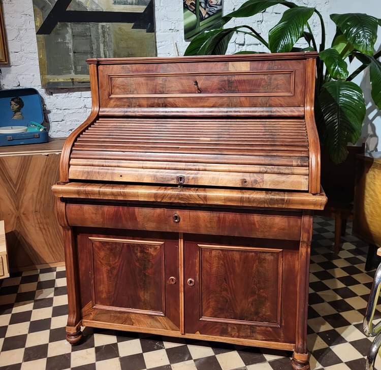 Antyczne biurko z roletą z ciemnego drewna, zamykane na klucz, z dwiema szafkami na dole, na tle szachownicy i rośliny doniczkowej.