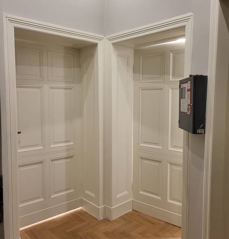 Dwa białe, symetryczne wejścia w ścianie, wykończone dekoracyjnymi listwami, z parkietem w jodełkę na podłodze i czarną skrzynką alarmową po prawej stronie.