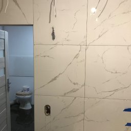 KRYSTIAN CHARCZENKO - Ściana łazienki w trakcie wykańczania, białe płytki imitujące marmur z niebieską taśmą malarską, widoczne przewody elektryczne, otwarta biała drzwi z szybą, w tle toaleta z wiadrem po farbie.