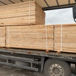 Prodrew Stanisław Dawiec - Stosy jasnej, świeżo obrobionej tarcicy, zabezpieczone białym sznurkiem, załadowane na pakę ciężarówki z widocznym kołem i fragmentem kabiny.