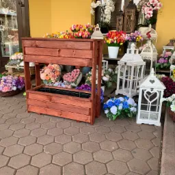 Drewniana, dwupoziomowa skrzynia na kwiaty z ciemnego drewna, otoczona kolorowymi bukietami kwiatów i białymi lampionami, ustawiona na brukowanym chodniku przed budynkiem z żółtą elewacją.