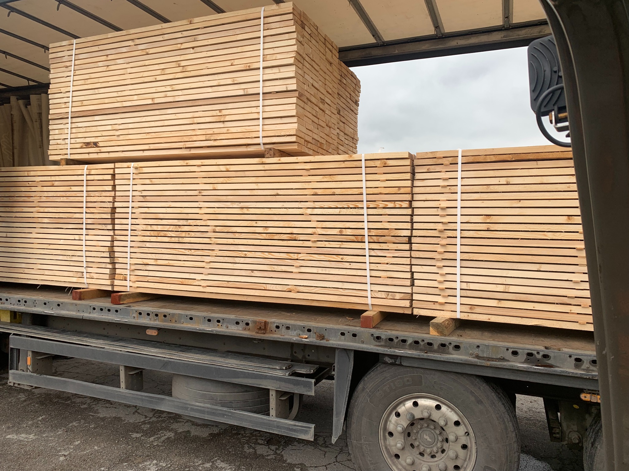 Stosy jasnej, świeżo obrobionej tarcicy, zabezpieczone białym sznurkiem, załadowane na pakę ciężarówki z widocznym kołem i fragmentem kabiny.