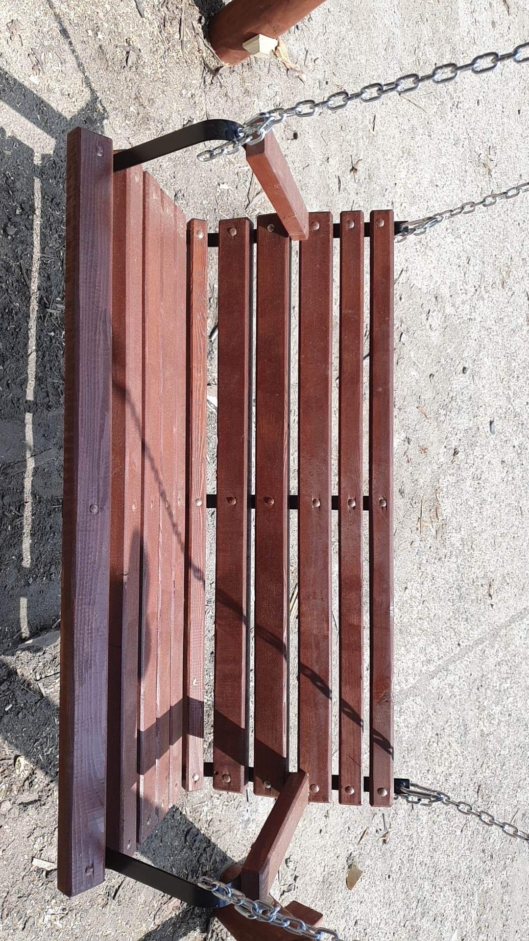 Widok z góry na huśtawkę z ciemnego drewna, zawieszoną na metalowych łańcuchach, rzucającą cień na szare podłoże.