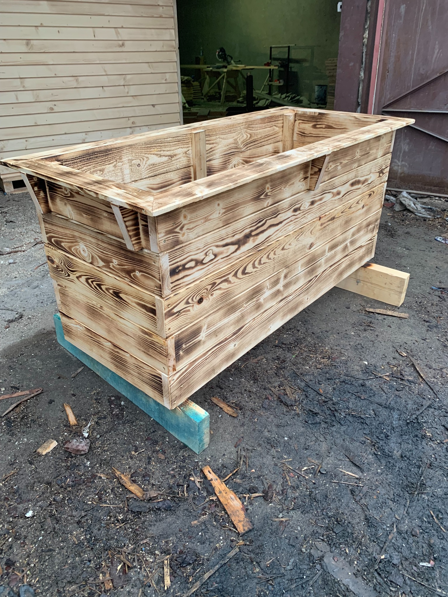 Drewniana skrzynia z opalonymi brzegami, stojąca na pomalowanych na niebiesko belkach, widoczna na tle drewnianej ściany i otwartych drzwi garażowych.