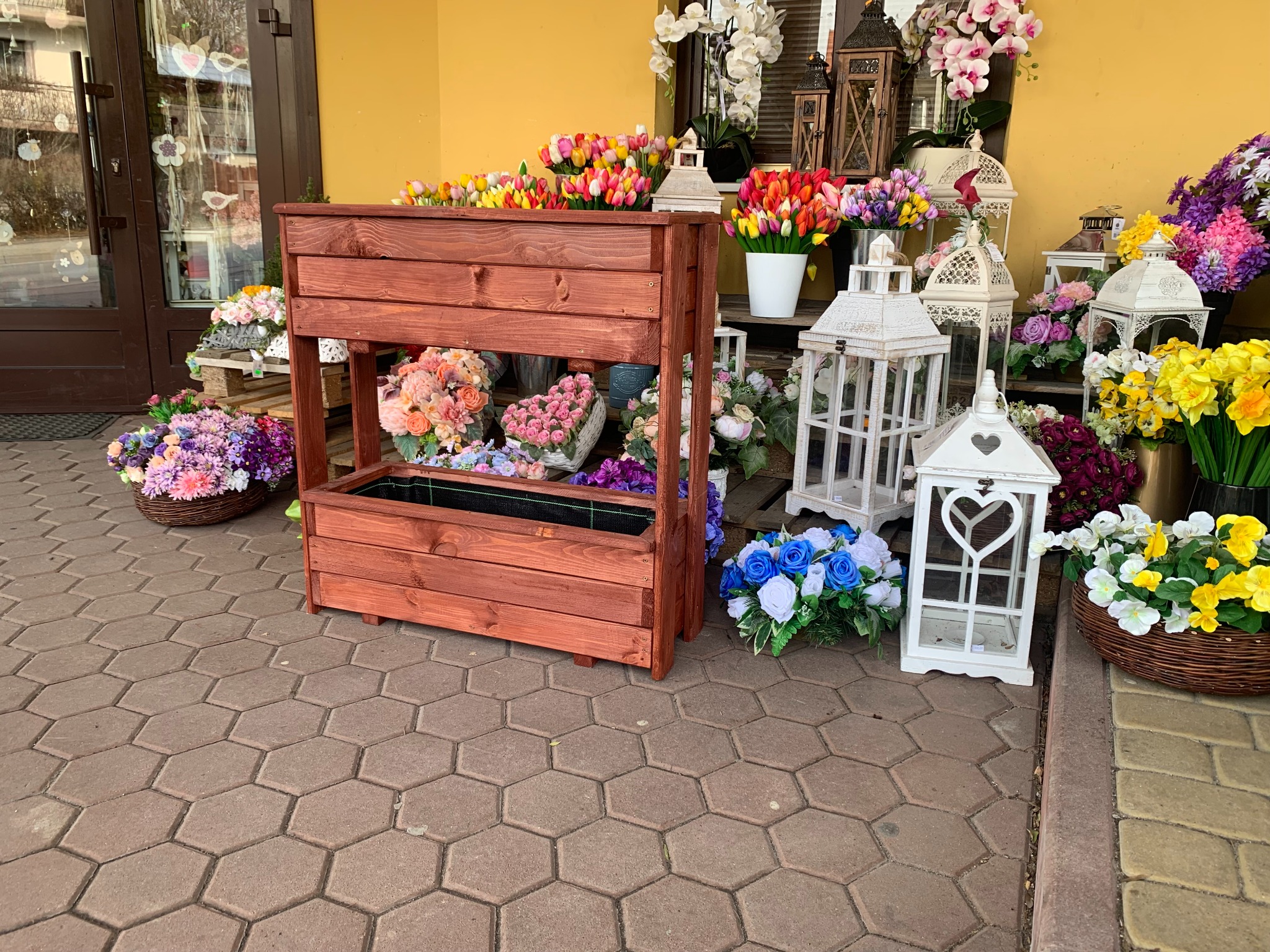 Drewniana, dwupoziomowa skrzynia na kwiaty z ciemnego drewna, otoczona kolorowymi bukietami kwiatów i białymi lampionami, ustawiona na brukowanym chodniku przed budynkiem z żółtą elewacją.