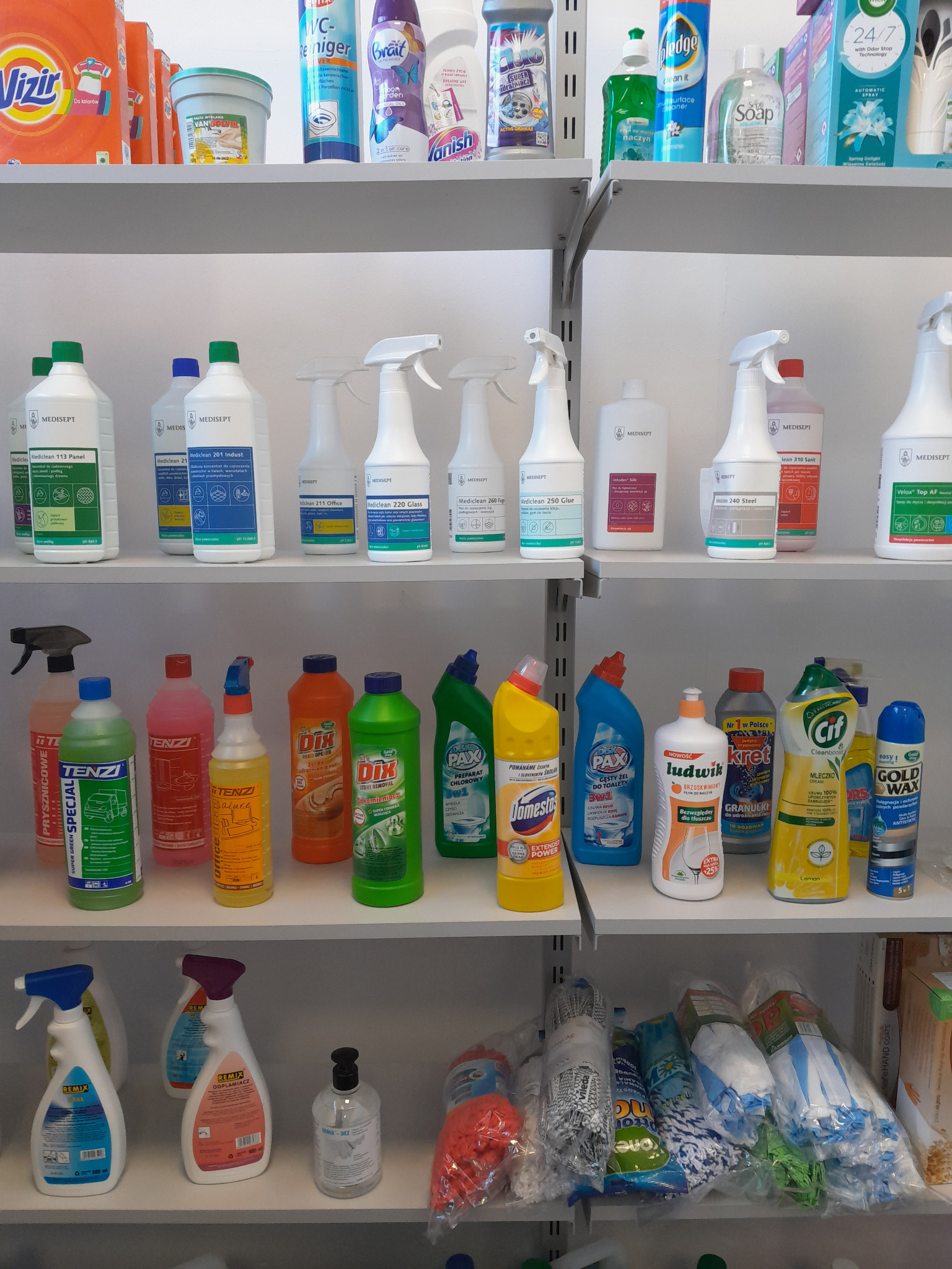 Półki sklepowe wypełnione różnorodnymi środkami czystości w kolorowych opakowaniach, w tym płyny uniwersalne, do toalet, do naczyń, oraz mopy i ściereczki w foliowych opakowaniach.