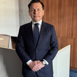 Kancelaria Adwokacka adwokat Piotr Bańczyk - Portret mężczyzny w ciemnym garniturze, białej koszuli i krawacie, stojącego w kancelarii z napisem 'Kancelaria Adwokat Piotr Ba', z zegarkiem na lewym nadgarstku.