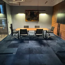 Nowoczesna sala konferencyjna z długim drewnianym stołem, krzesłami z siatkowym oparciem i abstrakcyjnym obrazem na ścianie, oświetlona dwoma lampami.