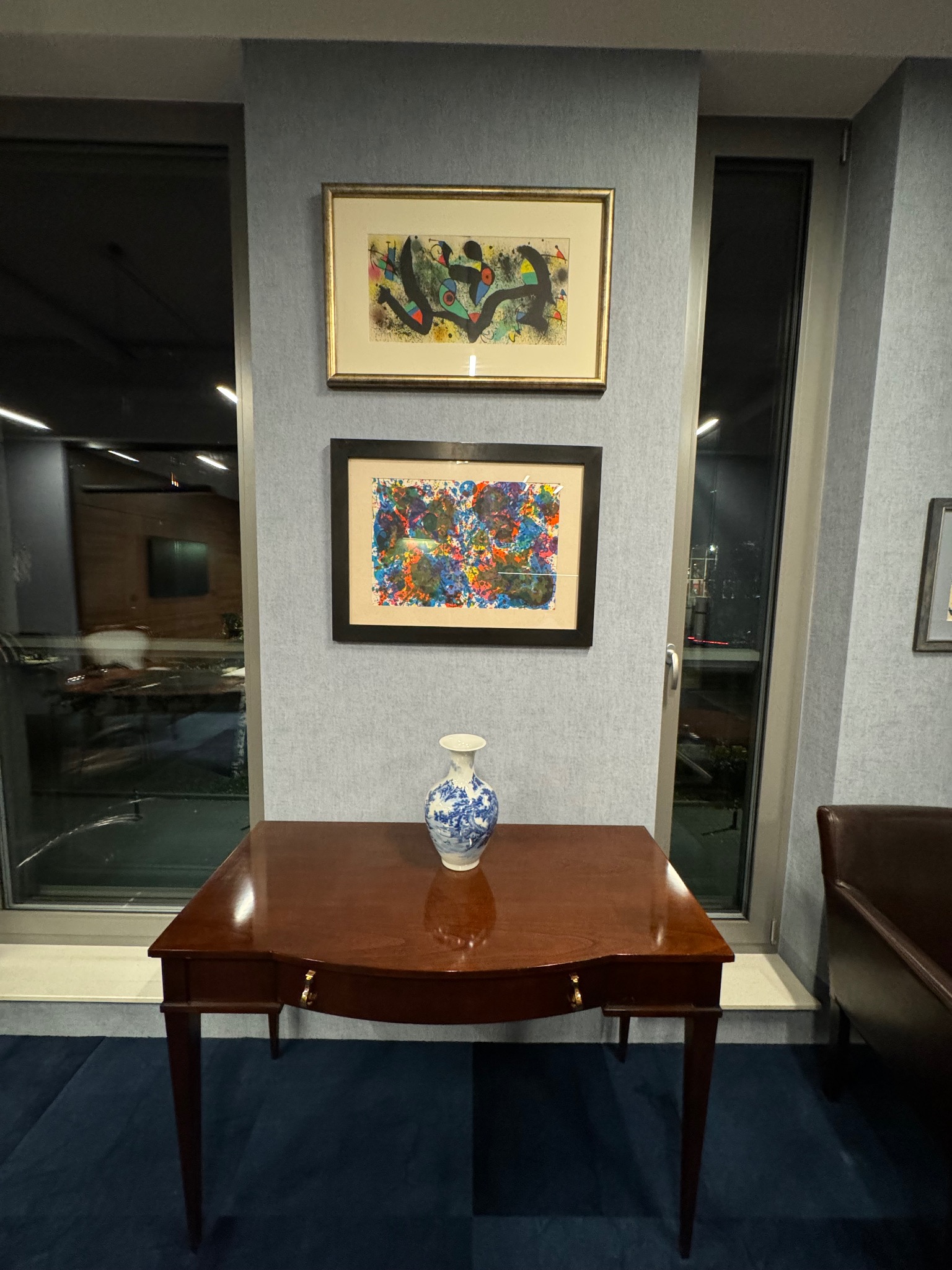 Eleganckie biuro z drewnianym biurkiem, na którym stoi wazon w niebieskie wzory, oraz dwoma abstrakcyjnymi obrazami na ścianie w tle.