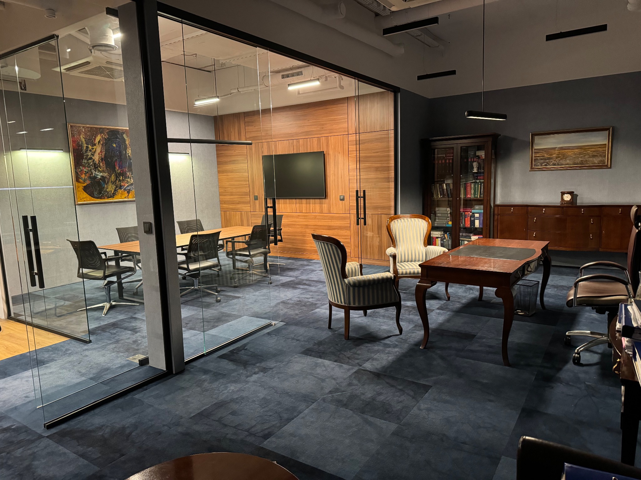 Przestronne, nowocześnie urządzone biuro z przeszklonymi ścianami, wyposażone w drewniane panele, duży telewizor, stół konferencyjny z krzesłami, klasyczne biurko i biblioteczkę, oświetlone...