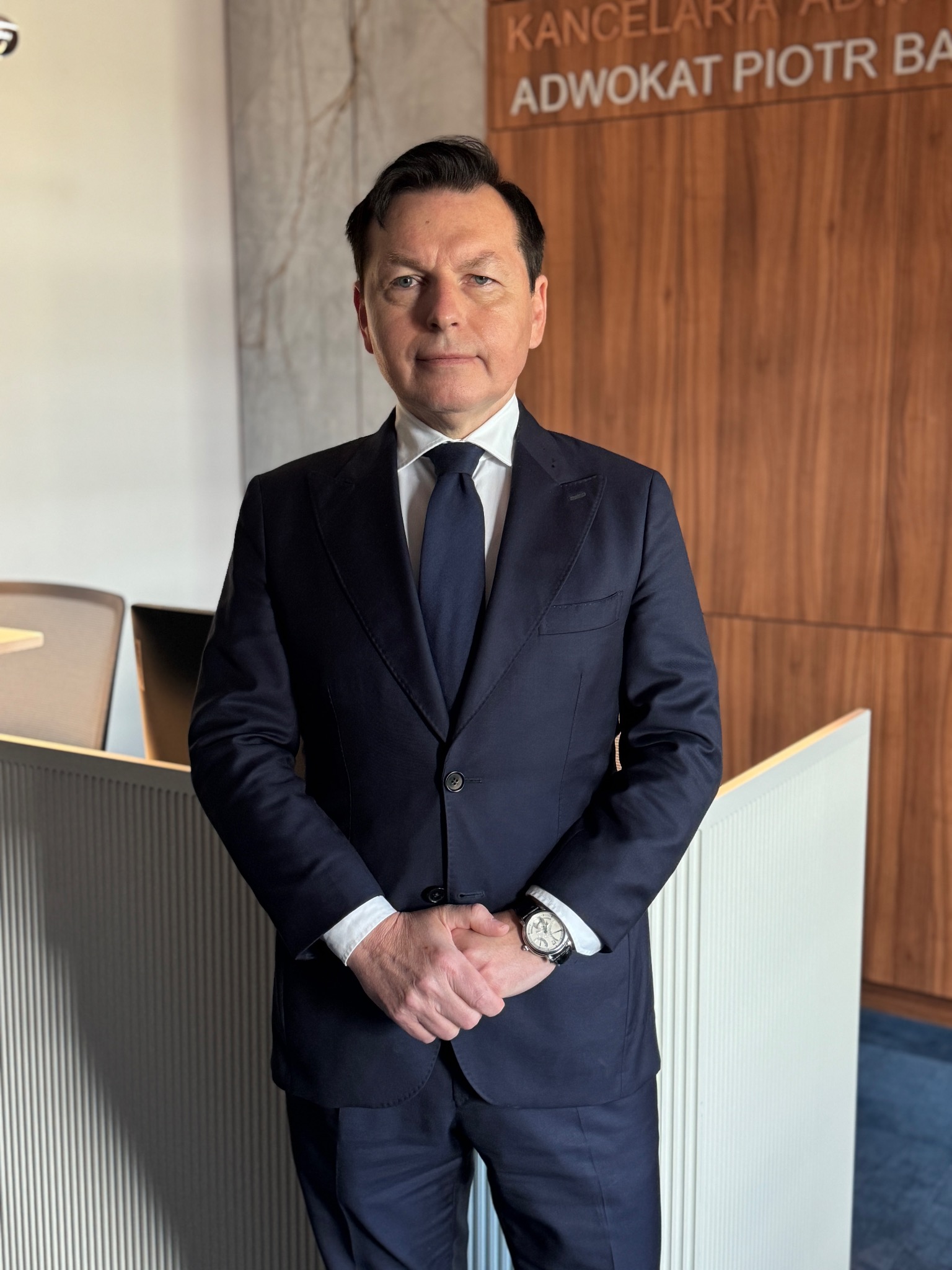 Portret mężczyzny w ciemnym garniturze, białej koszuli i krawacie, stojącego w kancelarii z napisem 'Kancelaria Adwokat Piotr Ba', z zegarkiem na lewym nadgarstku.