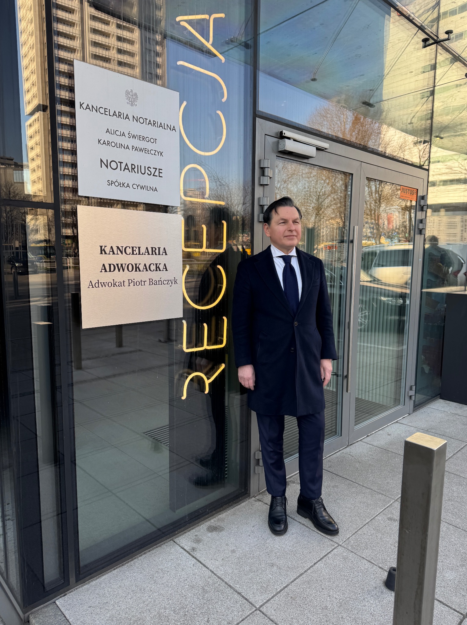 Mężczyzna w ciemnym płaszczu stoi przed nowoczesnym budynkiem z przeszklonym wejściem, obok tablic informacyjnych kancelarii notarialnej i adwokackiej, z neonowym napisem 'Recepcja' widocznym w tle.