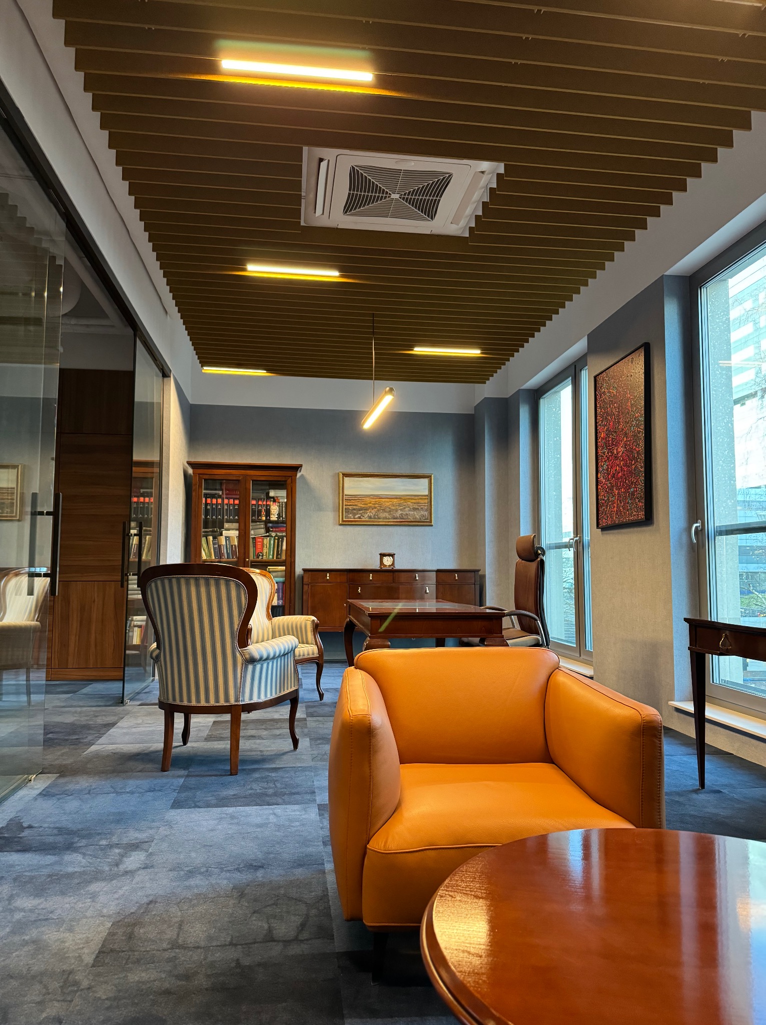 Nowoczesne biuro z elementami klasycznymi: pomarańczowy fotel, okrągły stolik kawowy, regał z książkami, obrazy na ścianach, sufit z oświetleniem liniowym.