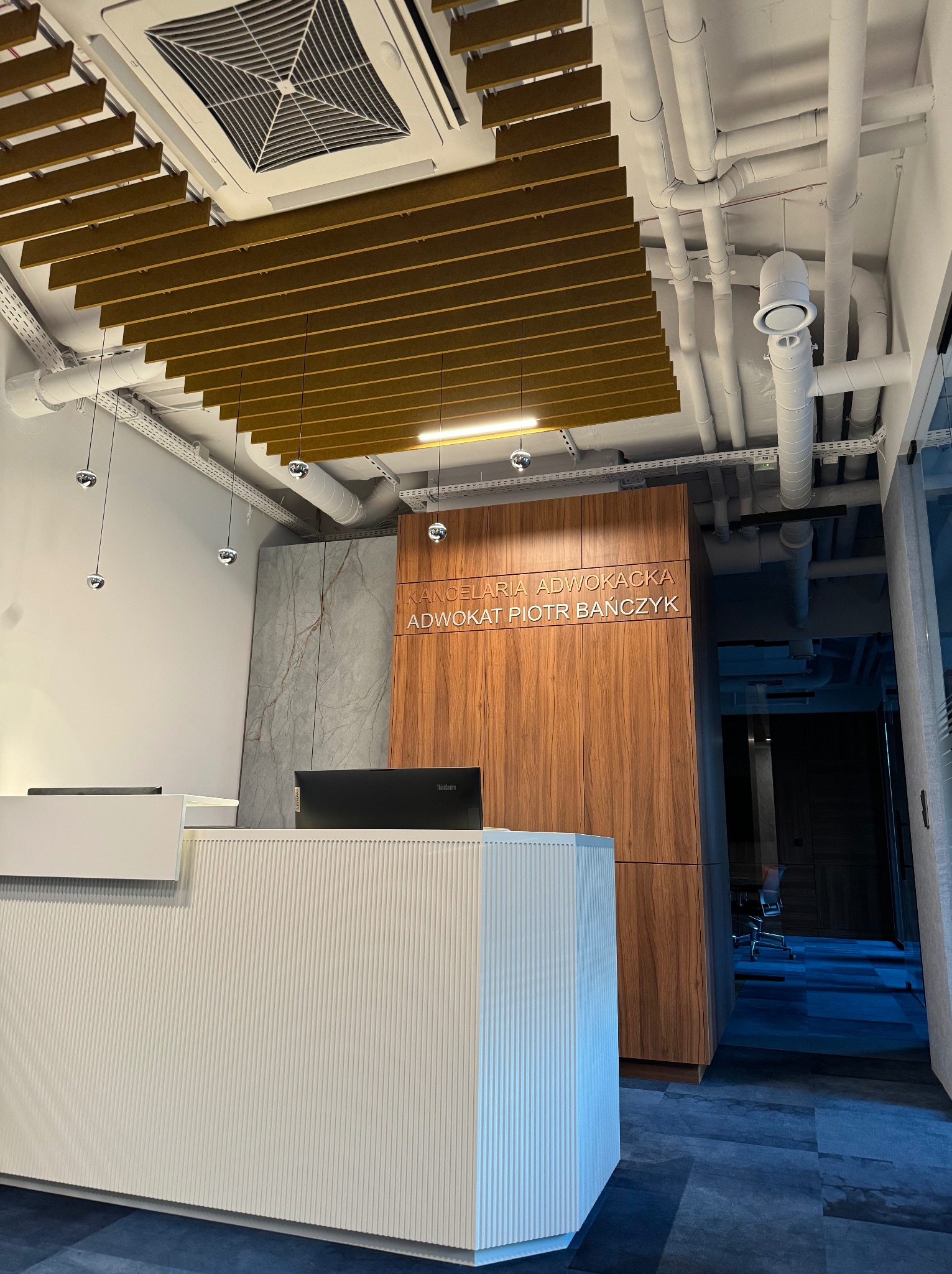 Recepcja w nowoczesnej kancelarii prawnej z białym biurkiem recepcyjnym o żłobkowanej fakturze, drewnianą ścianą z napisem 'Kancelaria Adwokacka Adwokat Piotr Bańczyk', designerskim oświetleniem...