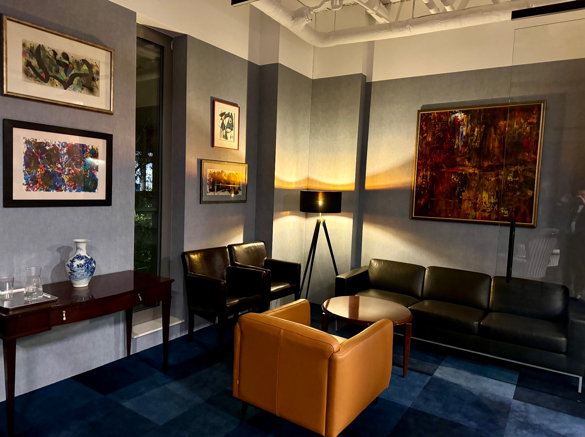 Eleganckie wnętrze biura z ciemnymi meblami skórzanymi, abstrakcyjnymi obrazami na ścianach, niebieską wykładziną i stołem z wazonem w stylu Delft.