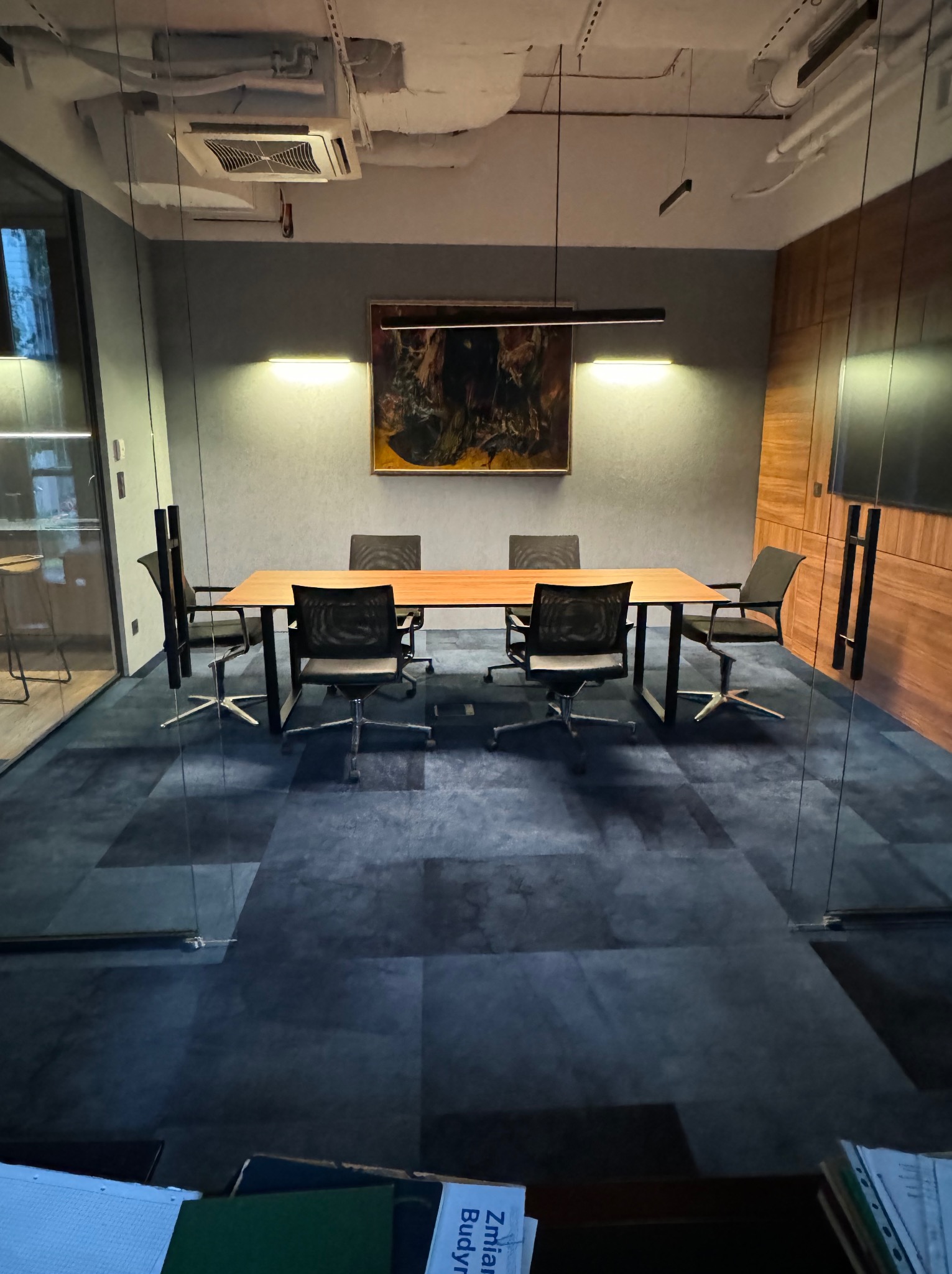 Nowoczesna sala konferencyjna z długim drewnianym stołem, krzesłami z siatkowym oparciem i abstrakcyjnym obrazem na ścianie, oświetlona dwoma lampami.