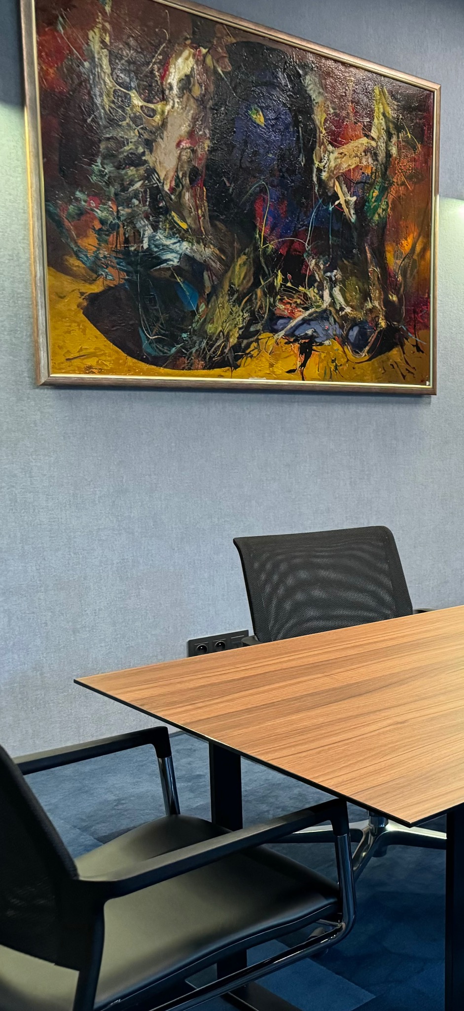 Nowoczesne biuro z drewnianym stołem konferencyjnym, czarnymi krzesłami z siatkowym oparciem i abstrakcyjnym obrazem w złotej ramie na szarej ścianie.