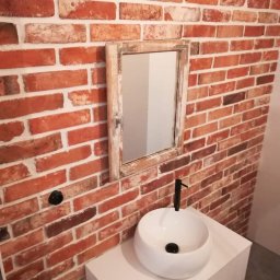 KAF-REM Wykończenia Wnętrz - Biała umywalka nablatowa z czarną armaturą na tle ściany z czerwonej cegły i lustra w postarzonej ramie.