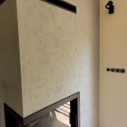 KAF-REM Wykończenia Wnętrz - Wykończony narożny kominek z czarną obudową i dekoracyjnym tynkiem na ścianie, widoczne gniazdka elektryczne i drewnopodobna podłoga ułożona w jodełkę.