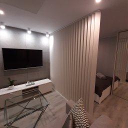 KAF-REM Wykończenia Wnętrz - Nowoczesne wnętrze: szara ściana z telewizorem, biała szafka RTV z rośliną, szklany stolik kawowy, ażurowa ścianka działowa oddzielająca sypialnię z widocznym łóżkiem i lustrzaną szafą.