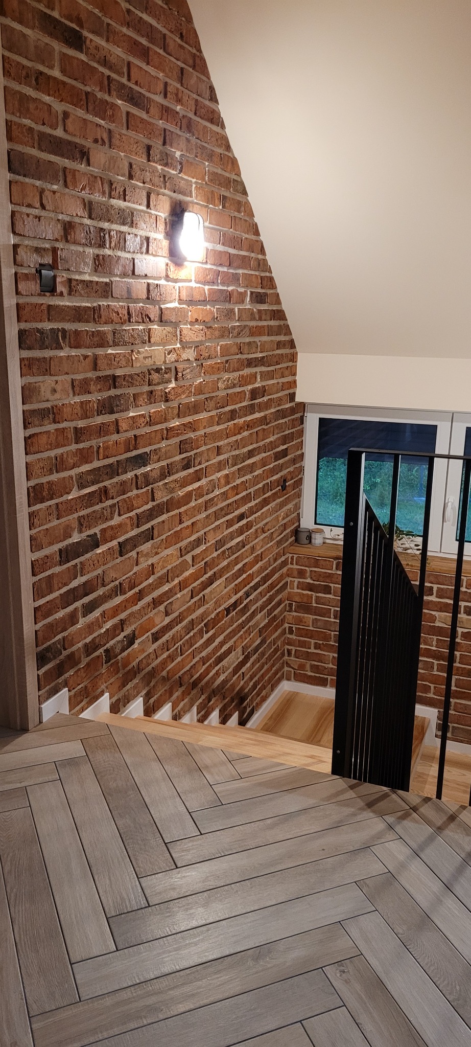 Ceglana ściana w klatce schodowej z oświetleniem punktowym i drewnopodobną podłogą ułożoną w jodełkę.