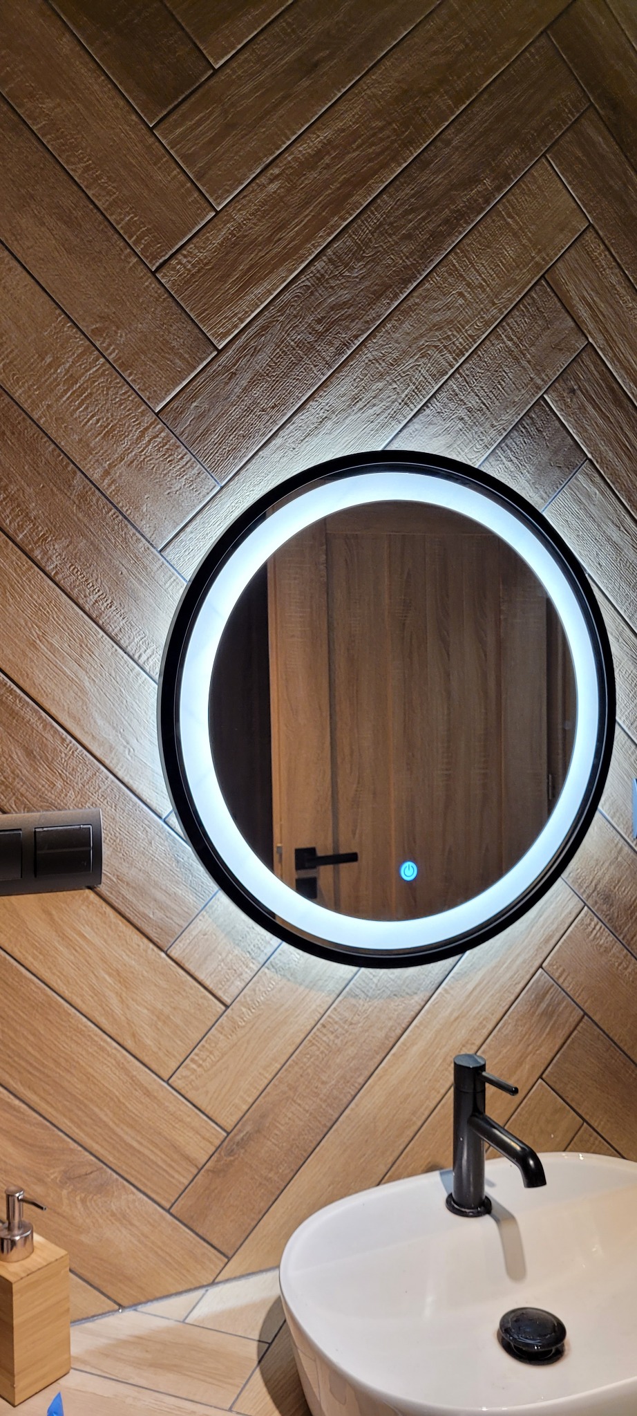 Okrągłe lustro z podświetleniem LED nad białą umywalką z czarną baterią na tle ściany wyłożonej drewnopodobnymi płytkami ułożonymi w jodełkę.