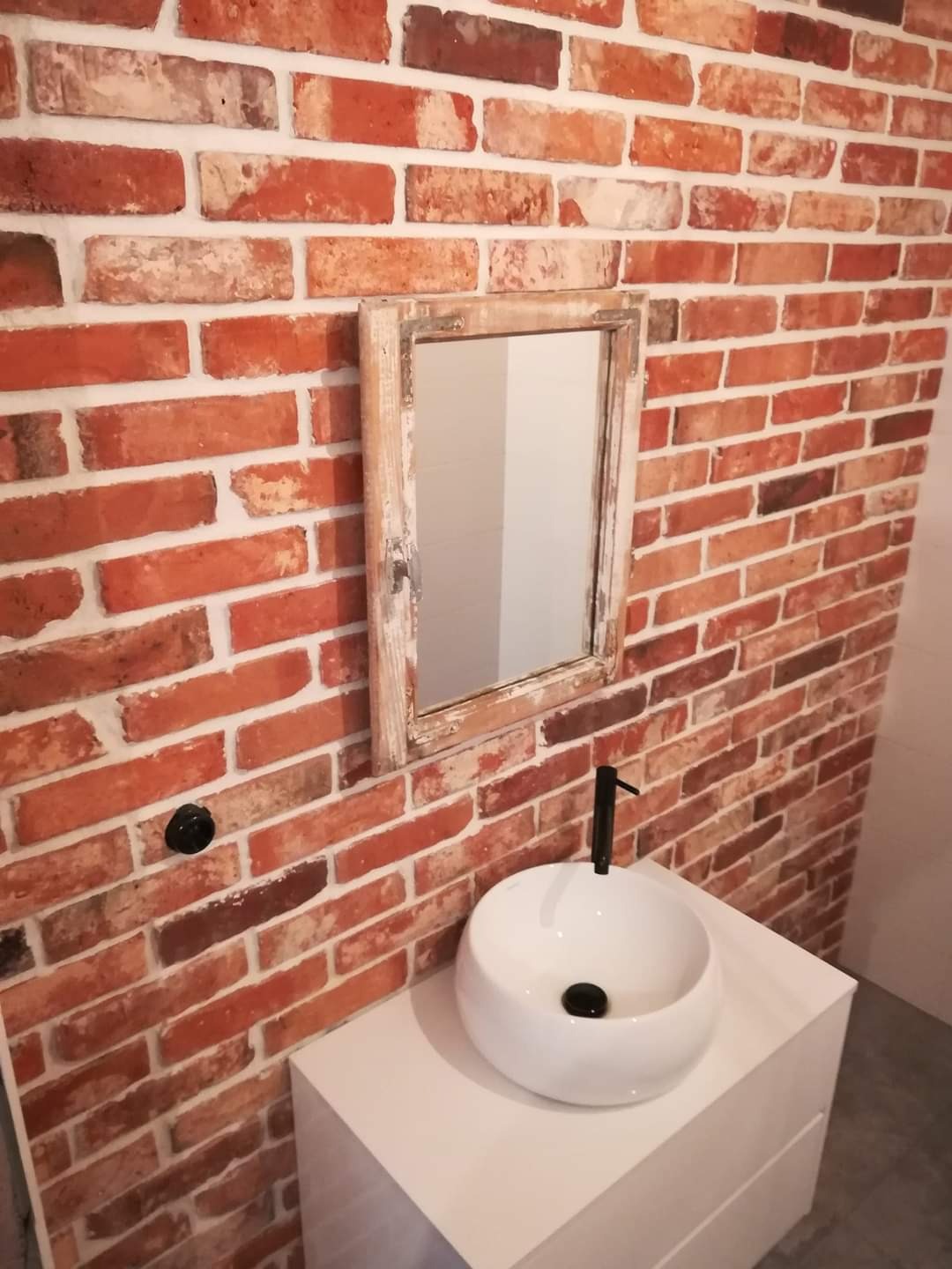 Biała umywalka nablatowa z czarną armaturą na tle ściany z czerwonej cegły i lustra w postarzonej ramie.