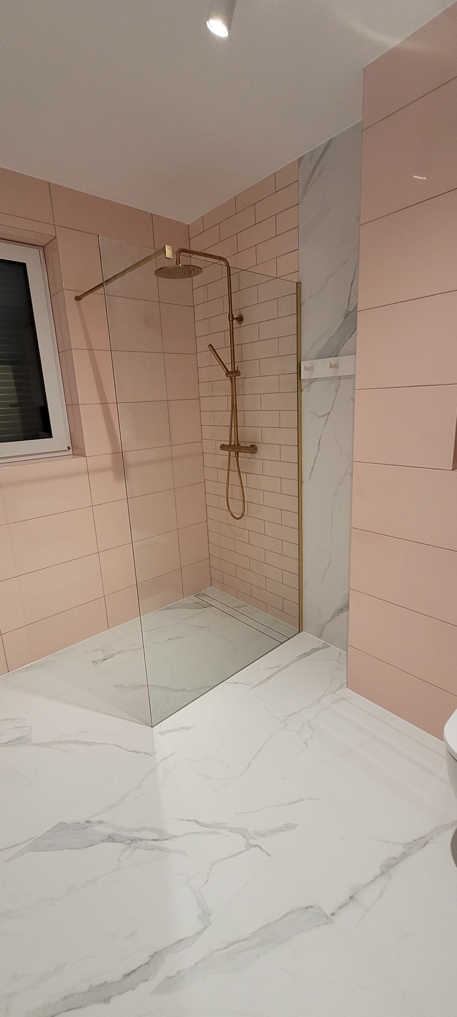 Nowoczesna łazienka z różowymi płytkami cegiełkami, marmurową ścianą, kabiną prysznicową typu walk-in ze złotą armaturą i białymi marmurowymi płytkami podłogowymi.