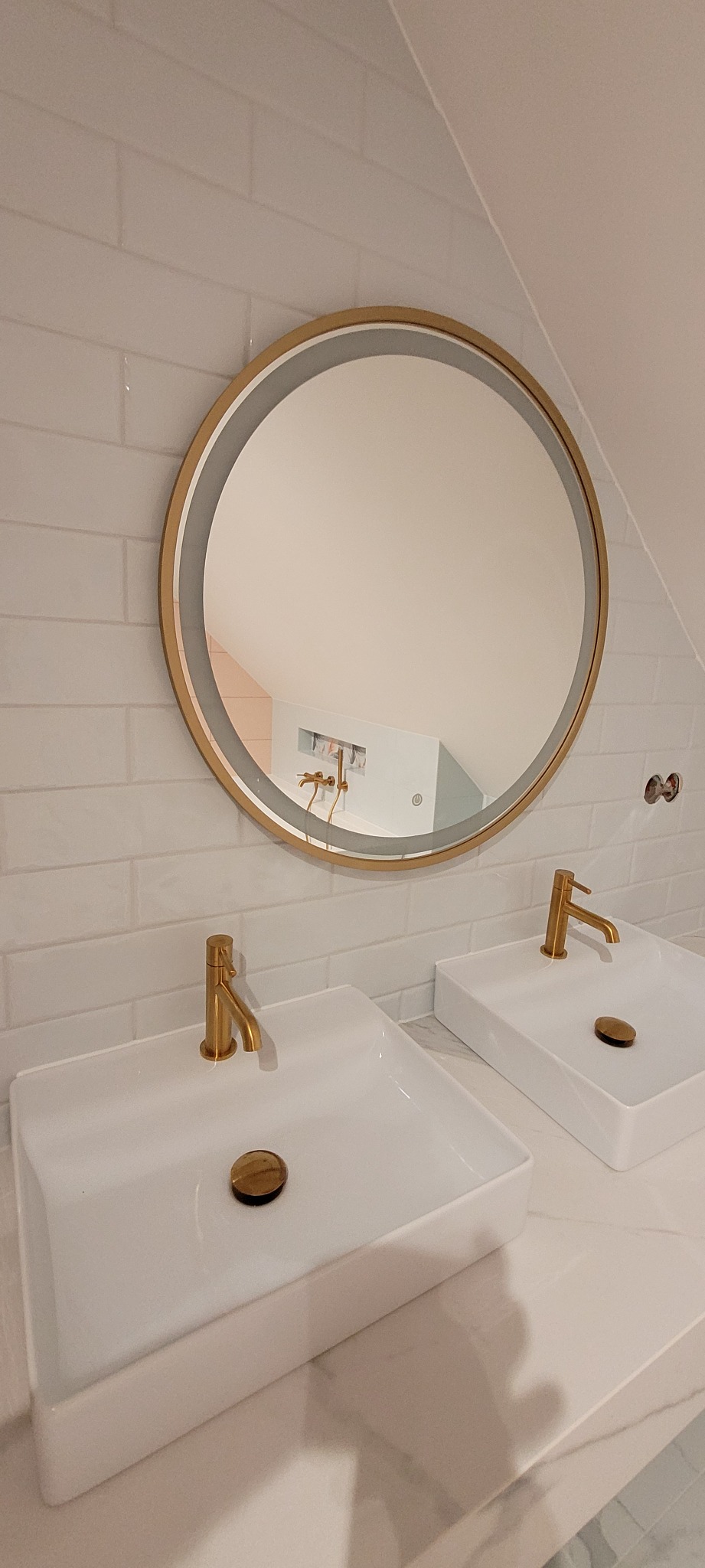Dwie białe, prostokątne umywalki z bateriami w kolorze złotym, okrągłe lustro z podświetleniem i złotą ramą, białe płytki na ścianie, widok na łazienkę odbity w lustrze.