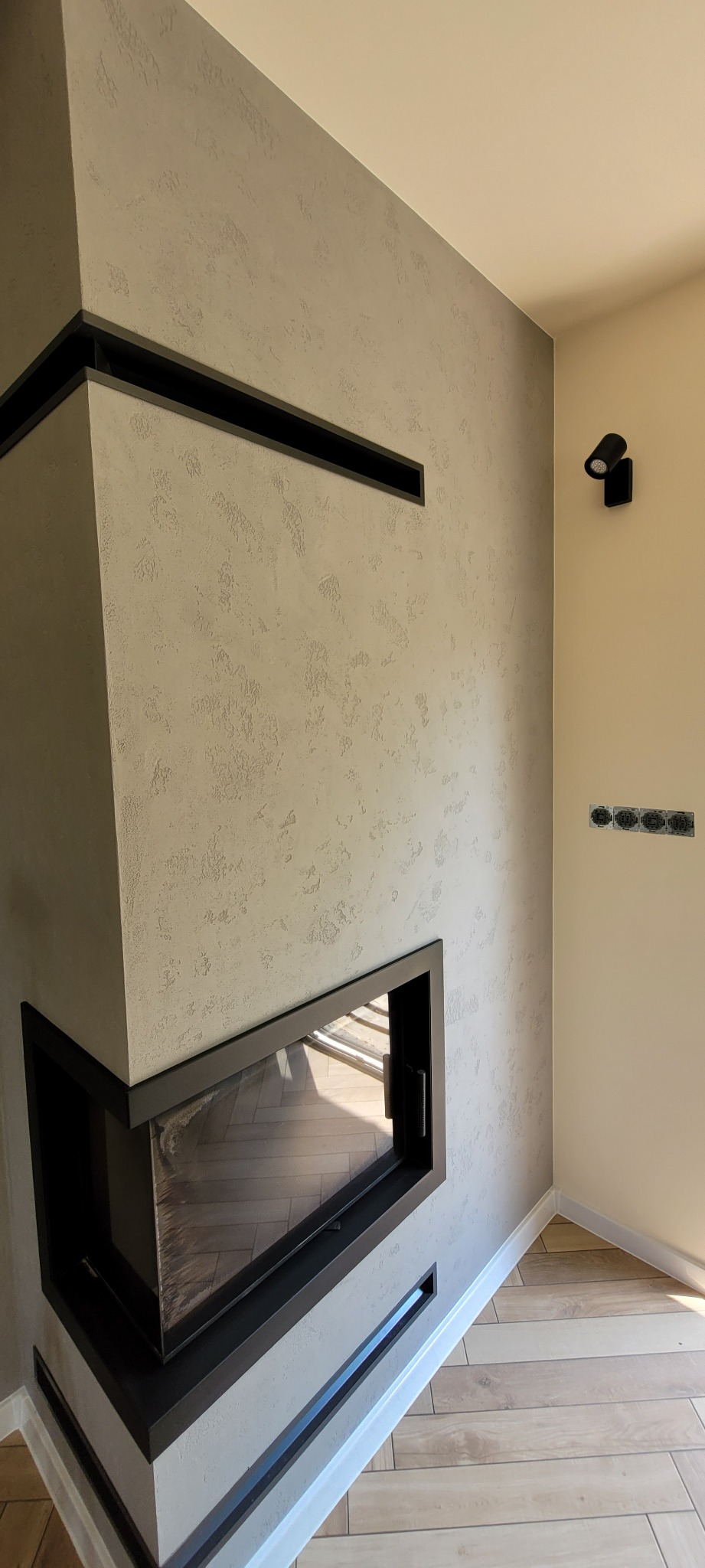 Wykończony narożny kominek z czarną obudową i dekoracyjnym tynkiem na ścianie, widoczne gniazdka elektryczne i drewnopodobna podłoga ułożona w jodełkę.