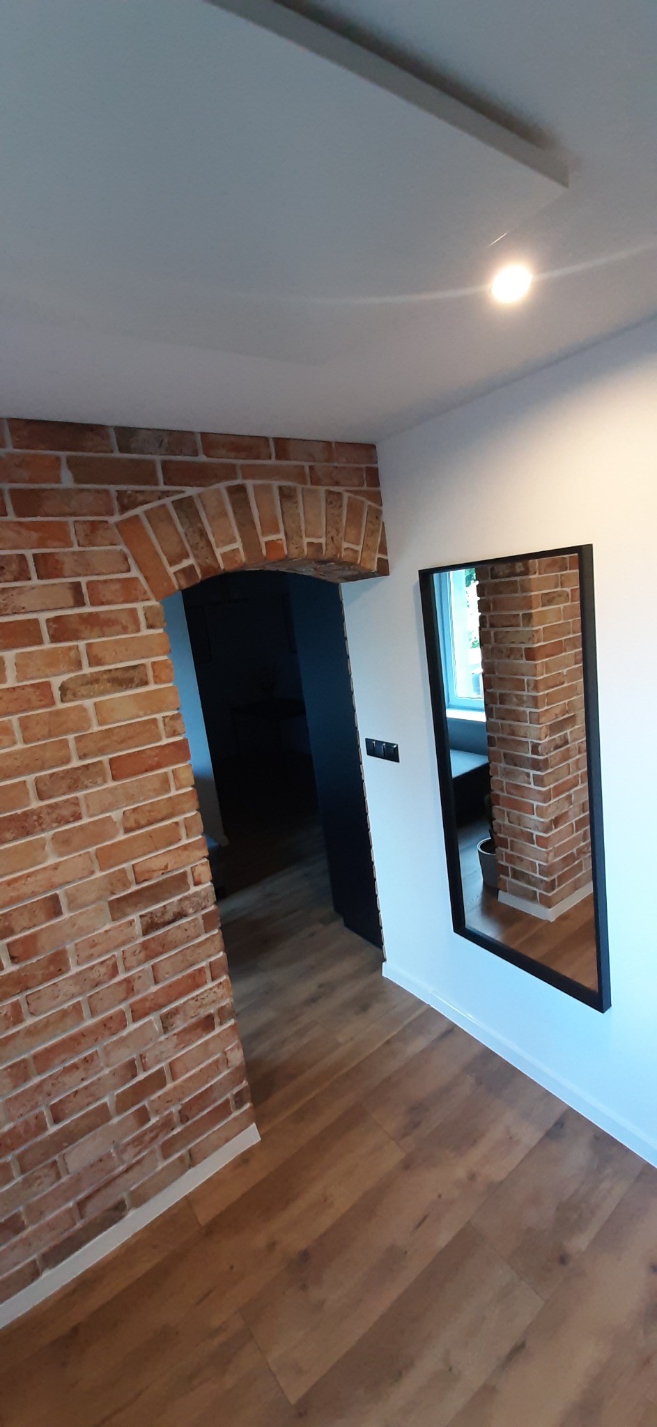 Wnętrze z ceglanym łukiem nad przejściem i lustrem odbijającym fragment ceglanej ściany, na podłodze panele drewnopodobne.