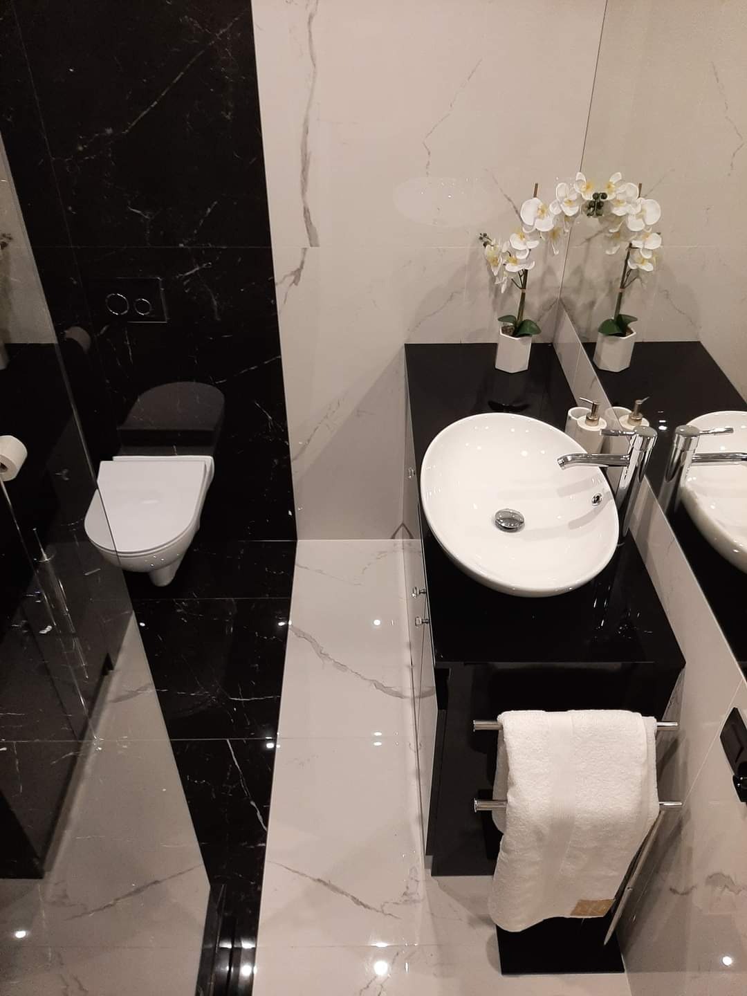 Nowoczesna łazienka z czarno-białymi marmurowymi płytkami, owalną umywalką na czarnym blacie, białym ręcznikiem na wieszaku i dekoracyjnymi białymi orchideami.