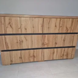 Komoda z sześcioma szufladami z drewnopodobnym frontem i czarnymi wstawkami, ustawiona na jasnej podłodze w minimalistycznym wnętrzu.