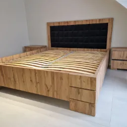 Drewniane łóżko z tapicerowanym, czarnym zagłówkiem w kwadraty, z drewnianymi szafkami nocnymi po bokach, bez materaca.