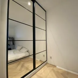 Nowoczesna szafa wnękowa z przesuwnymi drzwiami lustrzanymi i czarnymi ramami, odbijająca wnętrze sypialni z parkietem w jodełkę.